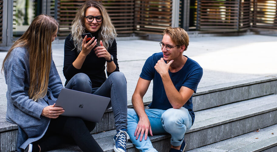 three young adults sitting on outdoor steps, engaged in conversation. One person is holding a laptop,